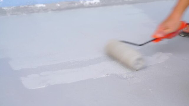 Worker applying grey waterproof coating to a flat surface with a paint roller. Concept of professional home renovation, roof sealing, and industrial floor maintenance. High quality texture.