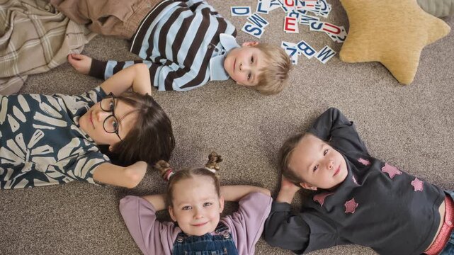 High angle portrait of joyful Caucasian children lying on floor in kindergarten playroom near star cushion and scattered alphabet flashcards and looking at camera