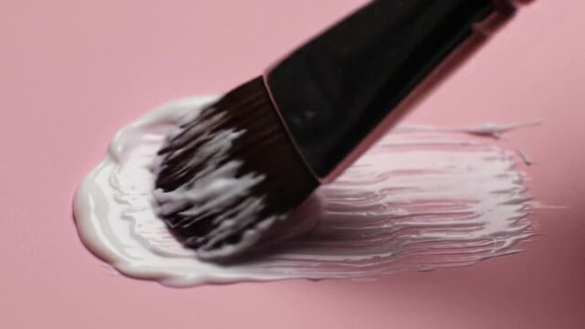 Close-up of a paintbrush creating a swathe of white cream on a soft pink surface.