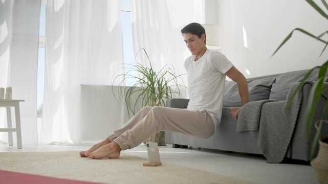 Athletic man exercising at home while performing triceps dips using a sofa in a bright modern living room. Young male, person or adult training barefoot near a smartphone, following an online workout