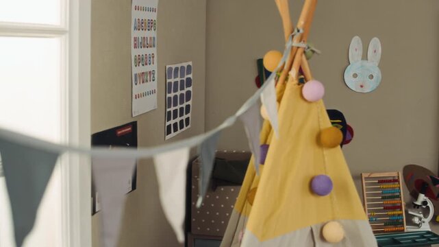 Tilt down no people shot of kindergarten classroom interior decorated with garlands, teepee tent, chalkboard with rules, weather chart and daily schedule on bright sunny day