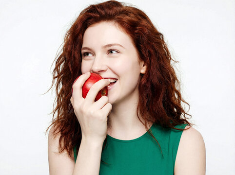 Redhead woman biting a fresh red apple and smiling against a white background.