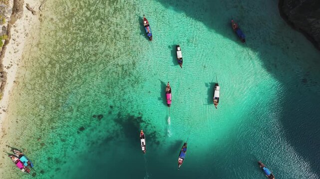Long tail boats drift through turquoise Maya Bay in Thailand, top down drone follow, slow motion shot.