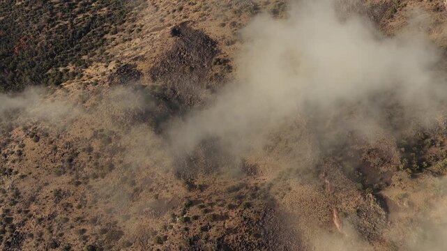 This cinematic drone shot captures a dramatic layer of morning mist and fog filling a deep, rocky desert canyon. The sun highlights the rugged textures of the volcanic rock and juniper landscape.