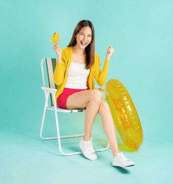 Happy smiling young Asian woman sit in deckchair and rubber ring in yellow color with holding credit card on blue background