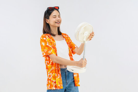 Happy smiling young Asian woman in summer wearing floral shirt holding fan to cool herself with suffers from too hot weather in white background