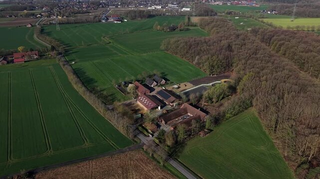 Aerial drone flyover of rural farmstead surrounded by green fields and forest during spring, with barns and buildings with solar panels in peaceful countryside landscape. Renewable energy production.