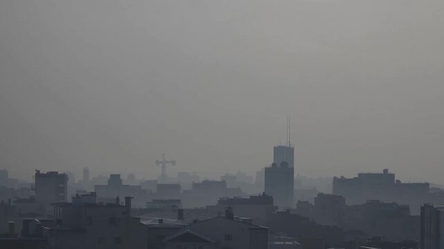 hazy city skyline at dawn, low contrast silhouettes of buildings and tower, cranes on horizon, dense smog and muted gray palette, industrial atmosphere and subdued light, slow panoramic movement