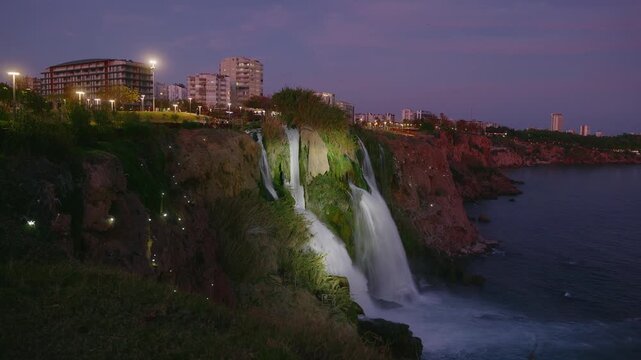 The Lower Duden Waterfall in Antalya, Turkey, tumbles over a cliff into the Mediterranean Sea at dusk. The cascade is beautifully illuminated against the twilight sky and the city lights