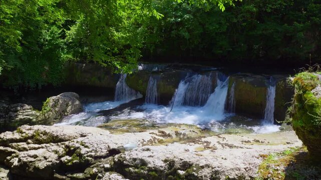 A beautiful waterfall cascades over mossy rocks in a pristine mountain river. Lush green trees surround the serene scene, with sunlight filtering through the leaves. A tranquil nature background