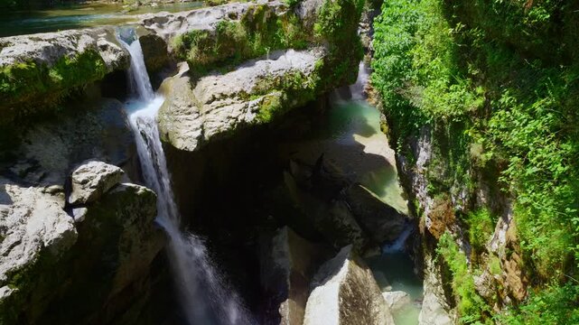 An aerial view of a powerful waterfall cascading into a deep rocky canyon. Lush green foliage covers the cliffs and turquoise water flows through the gorge on a sunny day
