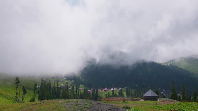 A serene view of Bakhmaro, a mountain resort in Georgia. Thick fog and clouds drift over green hills, pine forests, and a remote village nestled in a valley. A wooden yurt sits in the foreground