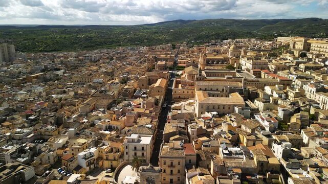 Aerial view of historic buildings and rooftops in Noto, Sicily, showcasing the intricate architecture and vibrant colors of the cityscape under a partly cloudy sky