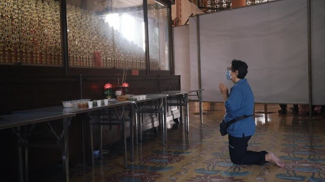Elderly Asian woman wearing a face mask kneels and prays with clasped hands in front of ancestral tablets and food offerings during the Qingming festival