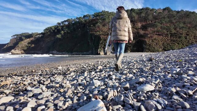 Woman standing on pebble beach facing ocean waves and cliffs