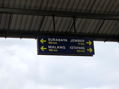 Overhead railway direction sign showing Surabaya, Jember, Malang, and Ketapang with distances in kilometers, mounted under metal roof with beams and sky visible.