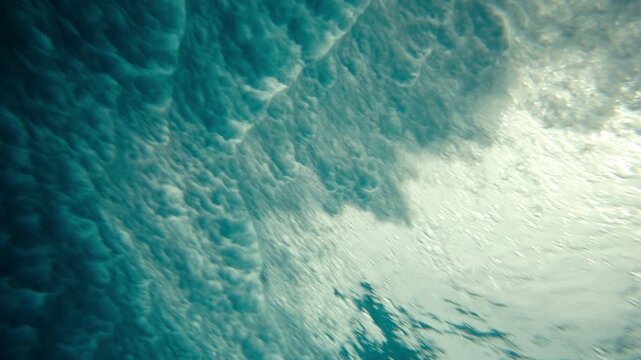 Underwater beneath large ocean wave showing textured blue water surface as whitewash crashes and bubbles foam and mist, backdrop background texture