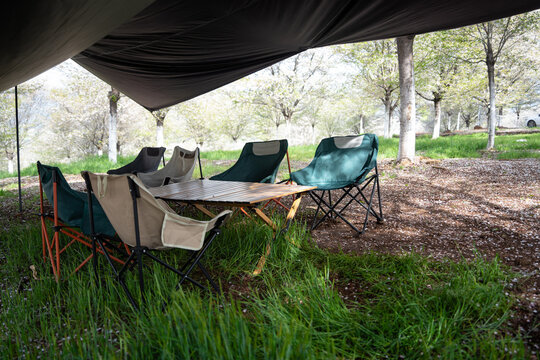 Outdoor camping setup with tarp shelter, folding chairs, and wooden table under trees in a grassy woodland area