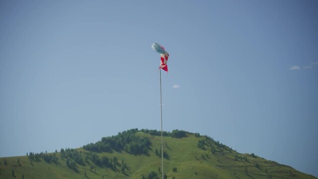 Windsock Fluttering Above Grassy Hilltop Under Clear Blue Sky, Tall Pole With Red And White Fabric Twisting In Strong Gusts, Wind Indicator For Paragliding Pilots And Meteorological Monitoring,