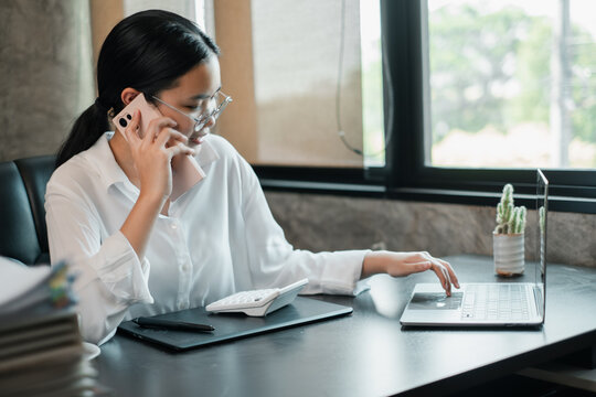 Professional woman in office using smartphone and laptop, multitasking efficiently in a bright, modern workspace.
