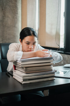 Focused young woman in a modern office, leaning on a stack of books, deep in thought, with glasses in hand.