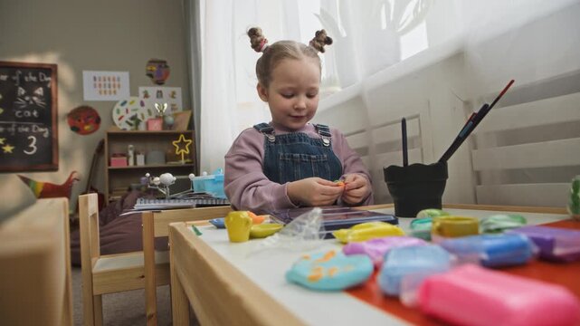 Medium handheld shot of Caucasian girl sitting at table and playing with modeling clay, focusing on creative activity in modern kindergarten classroom with boy playing in background