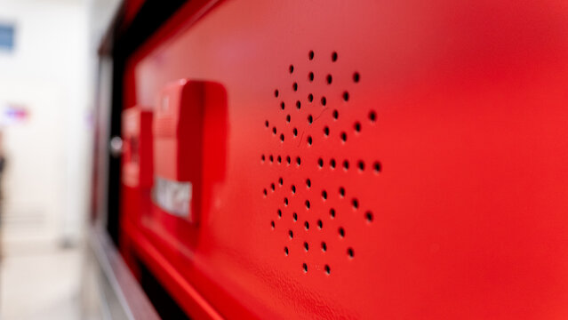 close up view of a red fire alarm control panel featuring a circular speaker grille and emergency notification components in a bright indoor environment