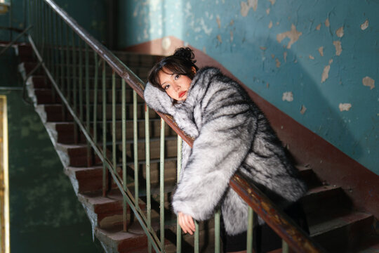 Woman in fur coat resting on railing in old staircase with blue walls