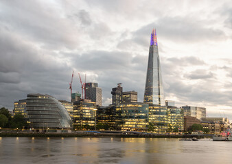 The London skyline rests beneath a heavy tapestry of twilight clouds, where the River Thames flows like liquid silk, mirroring the golden hum of the city. Dominating the horizon, the Shard pierces the