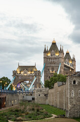 Fototapeta premium The northern Victorian Gothic tower and its adjacent arch stand bathed in warm, golden light, creating a sharp contrast against the cool, overcast sky. Vibrant blue suspension cables offer a striking 
