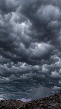 Dark storm clouds over rocky ridge, lightning flashes storm chaser documenting mammatus formations, rugged boulders in foreground, distant horizon glow, intense atmospheric drama