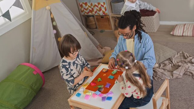 High angle handheld shot of young biracial male teacher engaging with Caucasian children sitting at table and playing with modeling clay, creating colorful figures during kindergarten art class