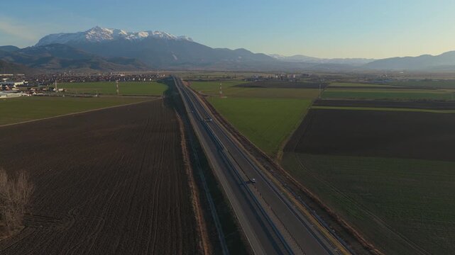 Epic new A3 highway with traffic crossing vast agricultural fields towards snowy Bucegi mountains near Rasnov Romania at golden hour, aerial lateral tracking right drone shot