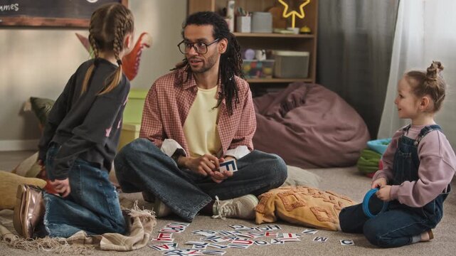 Ground level full shot of motivated young biracial male kindergarten worker teaching alphabet to Caucasian girls using flashcards, sitting on floor in cozy classroom and giving high five