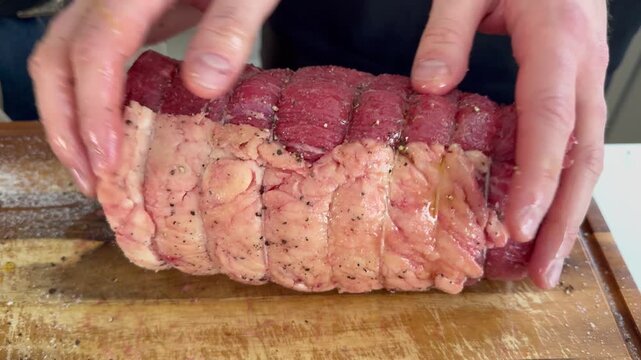 Closeup of a man&rsquo;s hands turning a raw topside joint of beef on a wooden board in a kitchen, while oil is being poured over and rubbed in to prepare the meat for roasting.