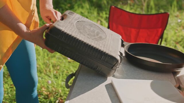 Woman outdoors setting portable stove, unpacking case and placing burner on table, connecting fuel and adjusting knob, hands checking latch and ignition, golden light in birch clearing ready for camp