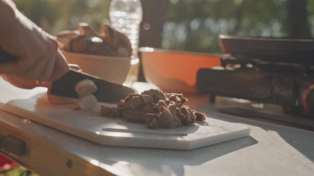 Caucasian hands slicing mushroom rounds outdoors at campsite, serene lakeside sunlight, cutting board with cooked meat and fresh mushrooms, portable stove and orange bowl suggest rustic saut and stew