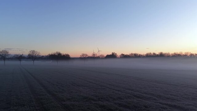 Aerial drone fly through misty farmland at sunrise in USA, with wind turbines emerging through fog in cold spring morning with soft golden light and atmospheric landscape. Dolly low forward.