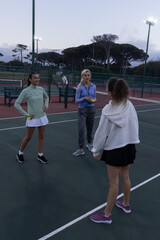 Obraz premium Female friends standing on fenced court at dusk holding yellow tennis balls near net in skirts