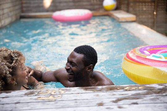 Couple leaning on pool deck and splashing water in backyard pool with rainbow-striped donut float