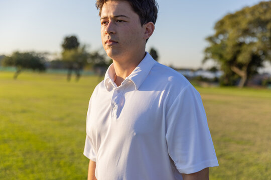 Man standing and looking right across grassy park during golden hour, wearing white polo shirt