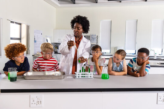 African American teacher guiding kids with goggles using pipette mixing green liquid at lab bench