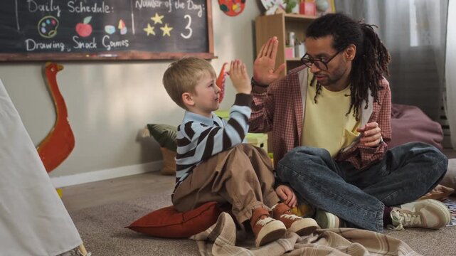 Handheld full slowmo shot of young biracial male teacher doing pinky swear and giving high five to little Caucasian boy sitting on floor in cozy kindergarten classroom