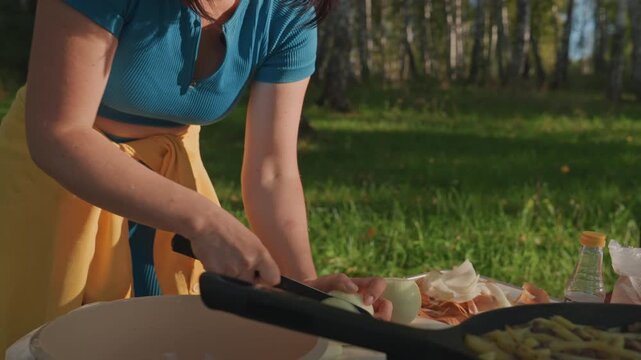 Woman frying breakfast on camp stove, skillet sizzling with eggs and bacon, tongs flipping food, closeup on hands and pan, warm morning light on grass, aromatic rustic outdoor meal