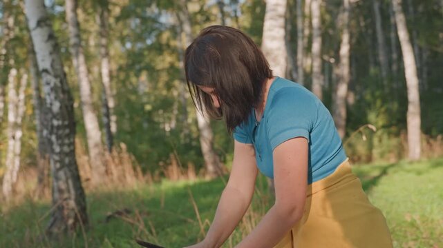 Woman setting portable stove forest camping table, birch grove backdrop and soft sunlight, car trunk visible in distance, arranging pan and utensils, preparing campsite kitchen for cooking, calm