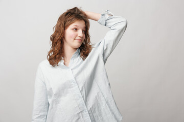 Naklejka premium Young woman with curly hair wearing a white shirt looks to the side with a slight smile, hand resting on her head. Neutral background, casual lifestyle and natural expression concept.