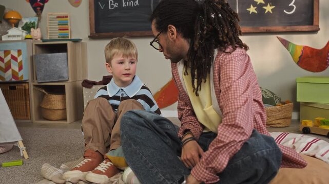 Handheld full shot of caring young biracial male teacher sitting on floor in cozy kindergarten classroom in front of blackboard and comforting upset Caucasian boy sharing his emotions