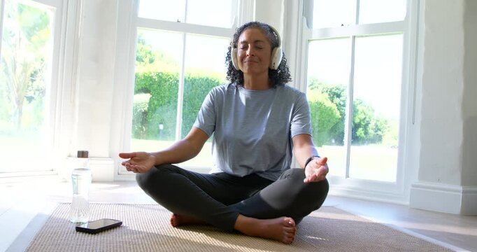 African American woman meditating on rug by bay window with headphones smartphone water bottle