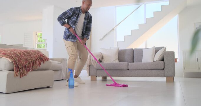 African American man entering picking mop spraying bottle mopping cleaning floor at home in plaid