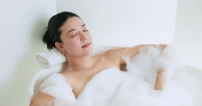 Noticing foam, woman moving arm through bubbles, leaning on rolled towel seeking calm in bathtub